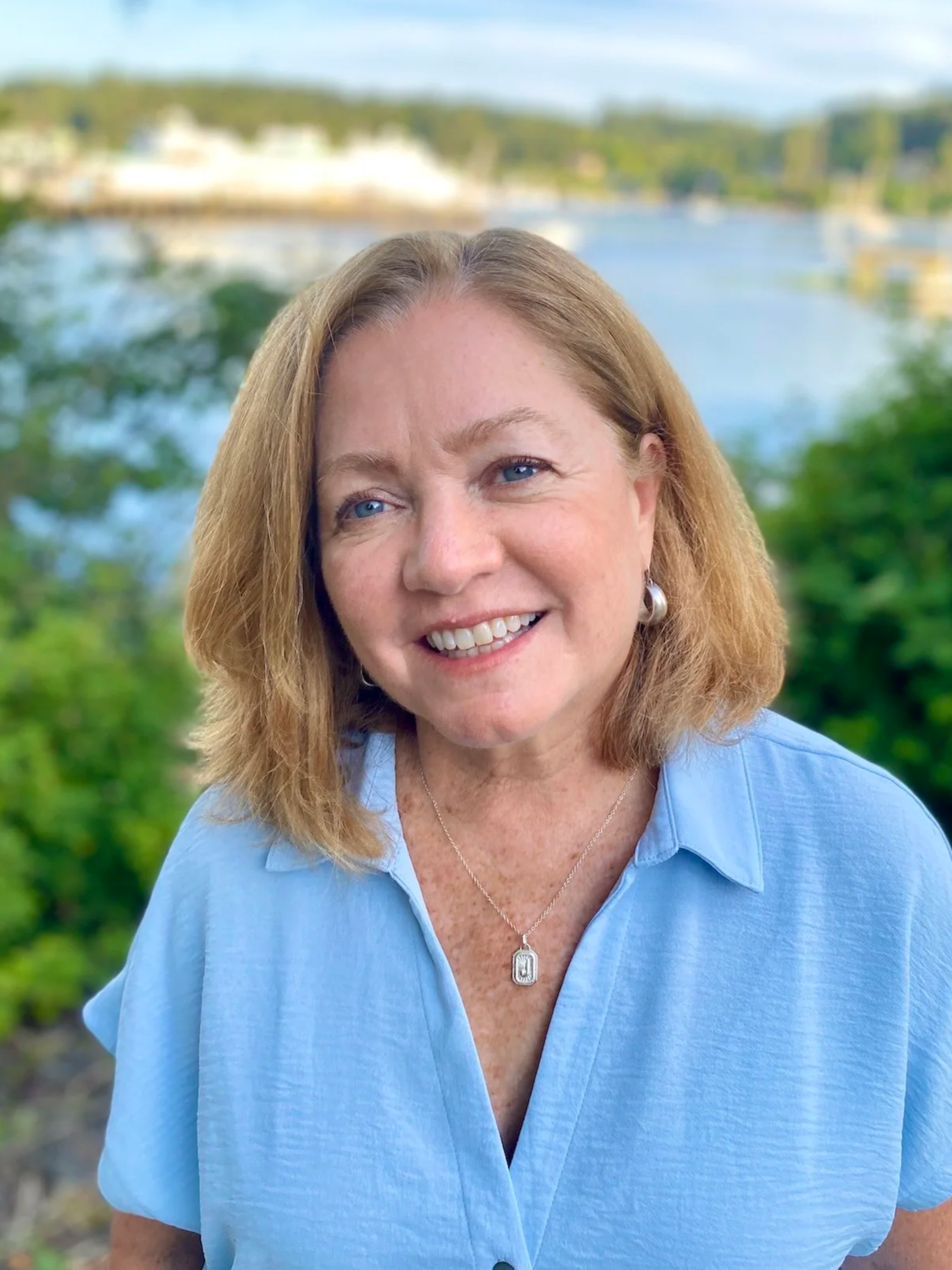 A smiling woman with shoulder-length blonde hair and blue eyes, wearing a light blue shirt, a silver necklace with a rectangular pendant, and pearl earrings, posing outdoors with a blurred background of greenery, water, and trees.