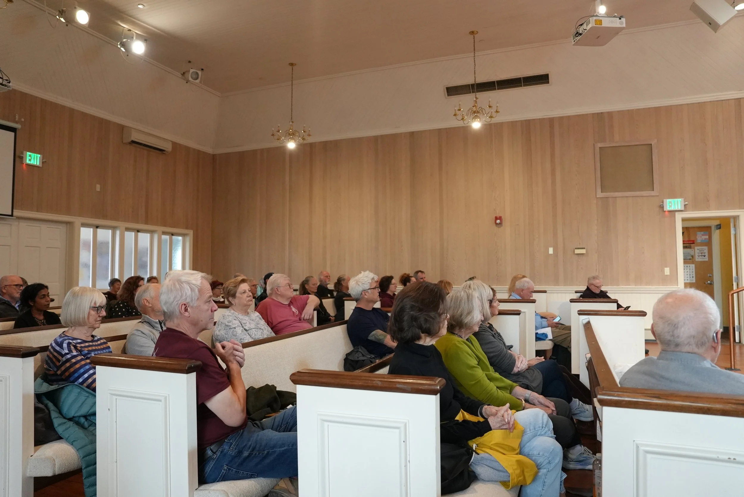 People seated in a church or meeting hall listening to a speaker, with wood-paneled walls, chandeliers, and emergency exit signs visible.