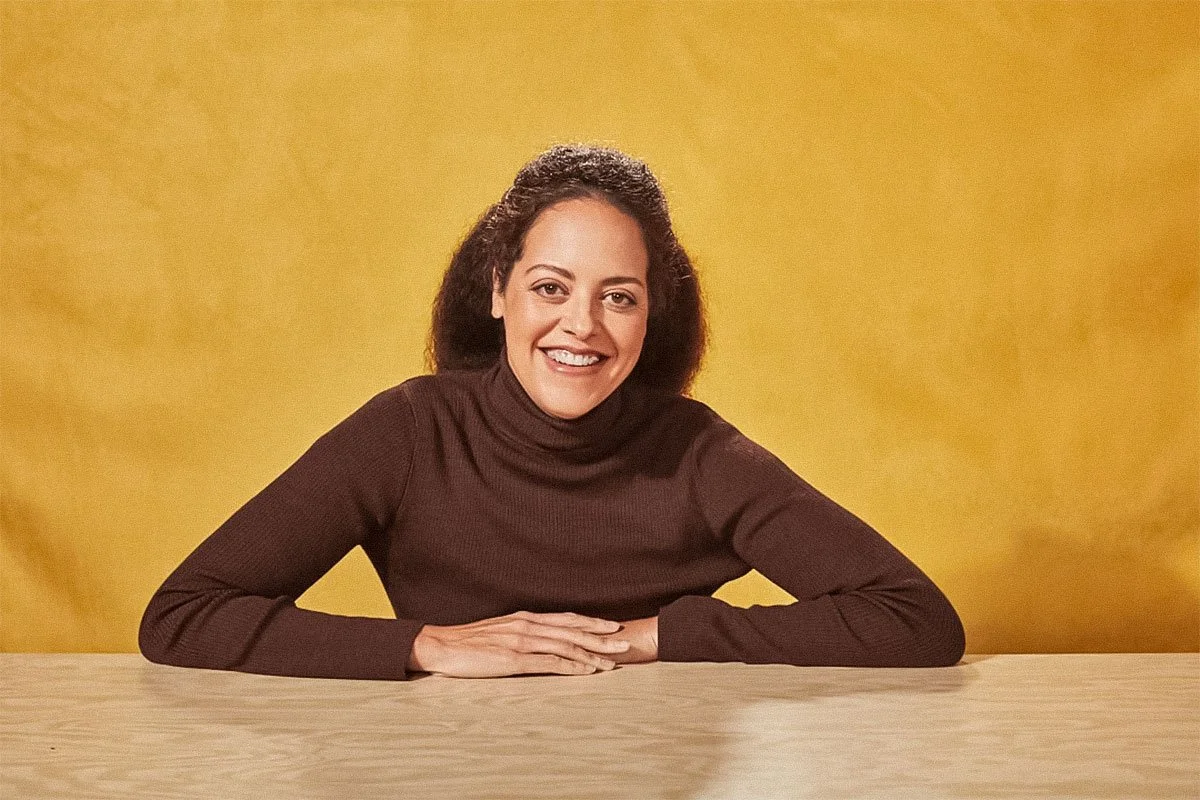 Woman with curly hair smiling, sitting at a wooden table, wearing a dark turtleneck sweater against a yellow background.