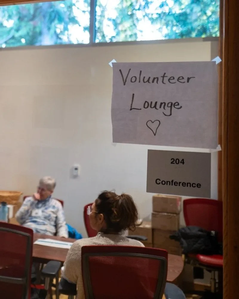 Sign for Volunteer Lounge with a heart drawing, located in a conference room with two women sitting at a table.