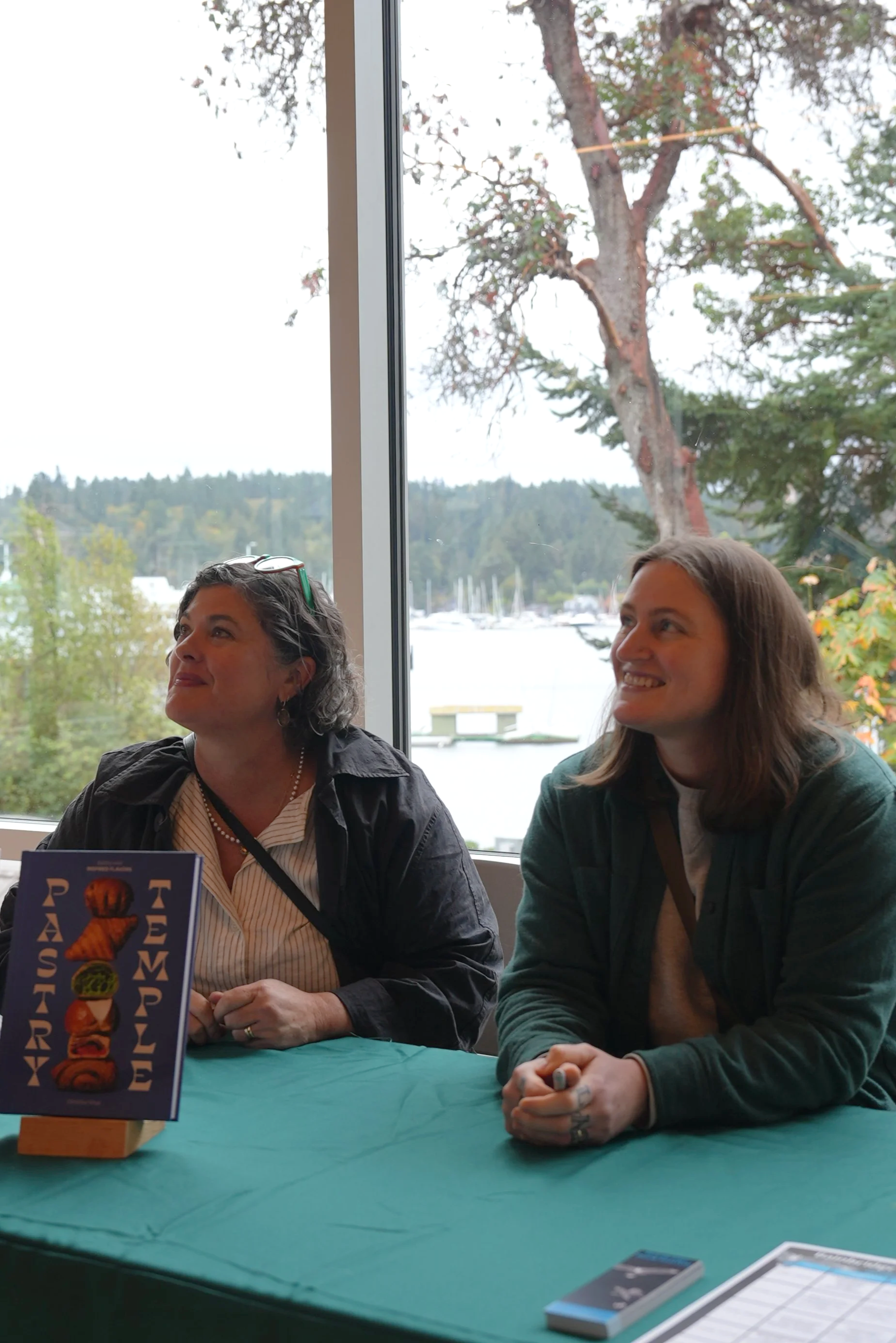Two women authors sitting at a book signing table near a window, smiling and looking away from the camera, with trees and a marina visible outside.
