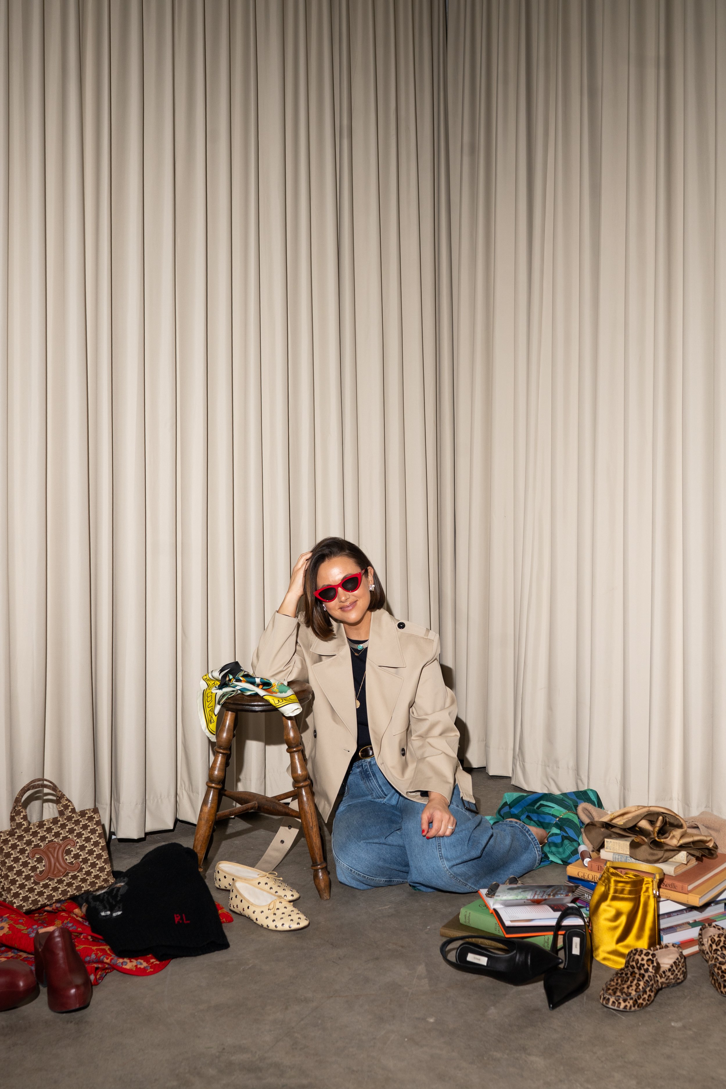 A woman with dark hair wearing red sunglasses, a beige trench coat, and blue jeans, sitting on the floor in front of a beige curtain, surrounded by fashion items and books, with one hand resting on her head and a slight smile.