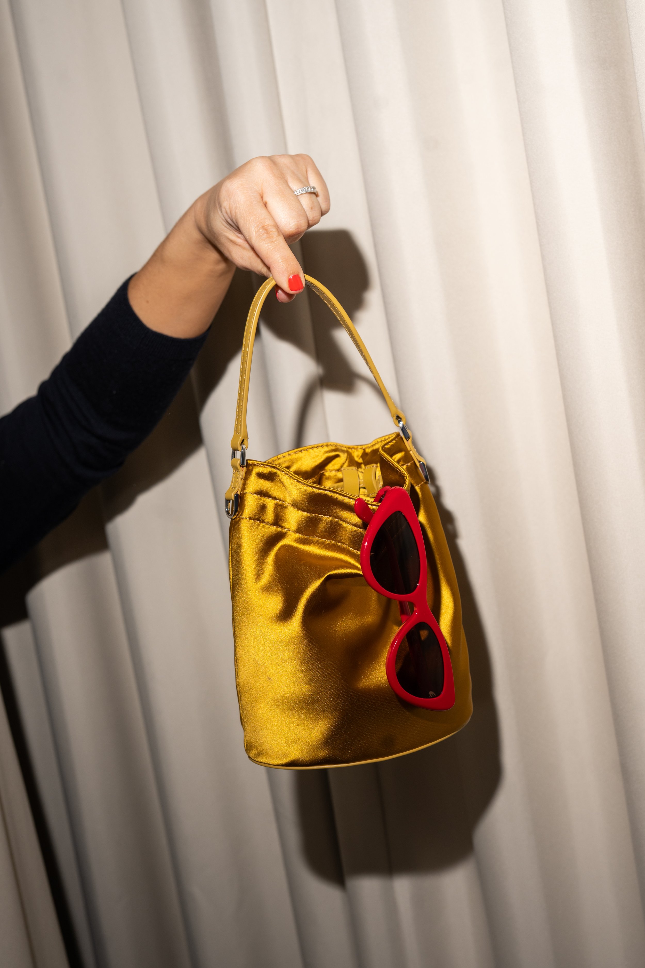 A person's hand holding a small, shiny yellow handbag with a red pair of sunglasses hanging from it, against a neutral curtain background.