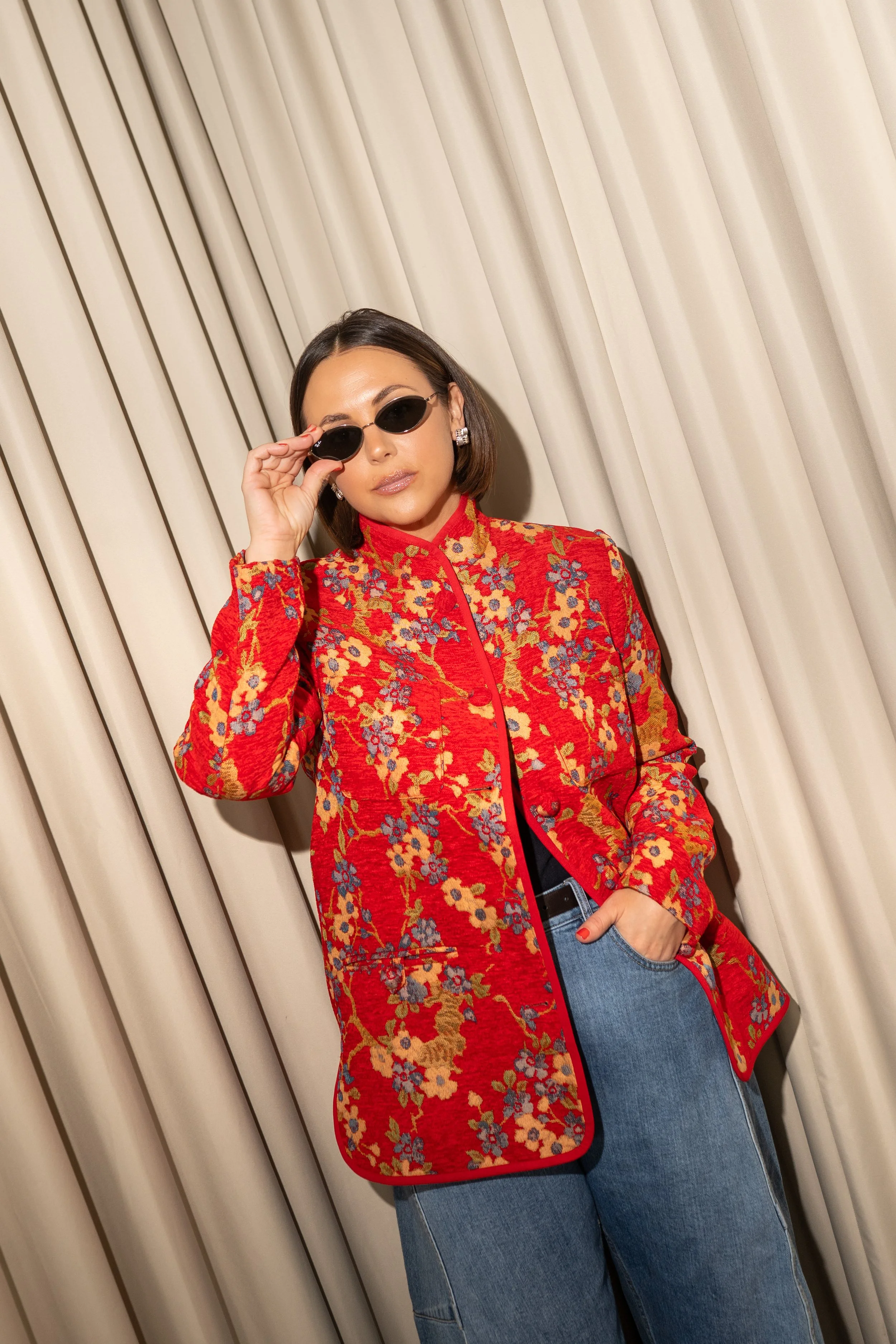 Woman wearing a red floral embroidered jacket, blue jeans, black sunglasses, and earrings, standing in front of beige curtains.