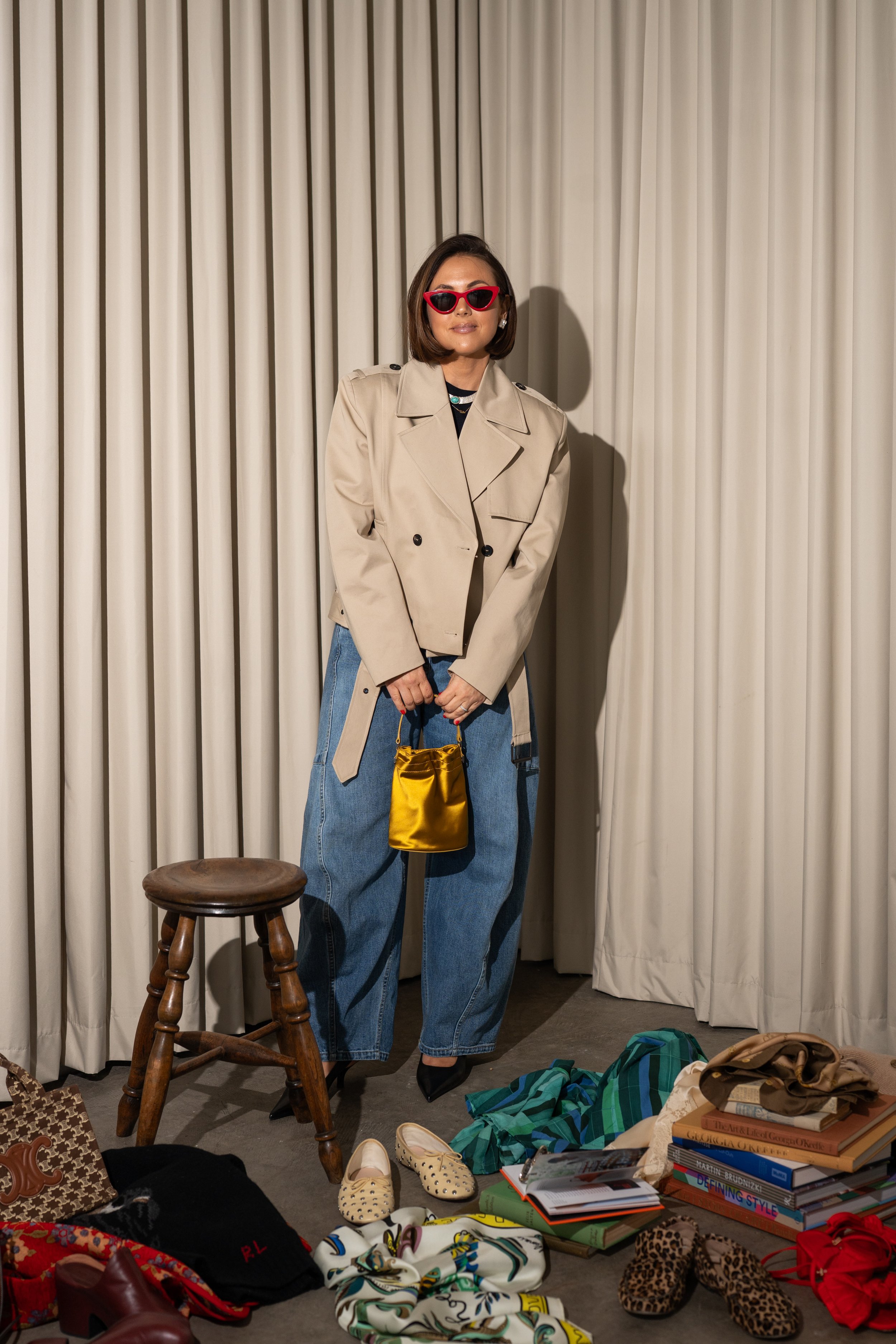 Woman in beige blazer, blue jeans, black heels, red sunglasses, holding a small yellow satin pouch, standing among shoes, books, and clothing on the floor in front of cream-colored curtains.