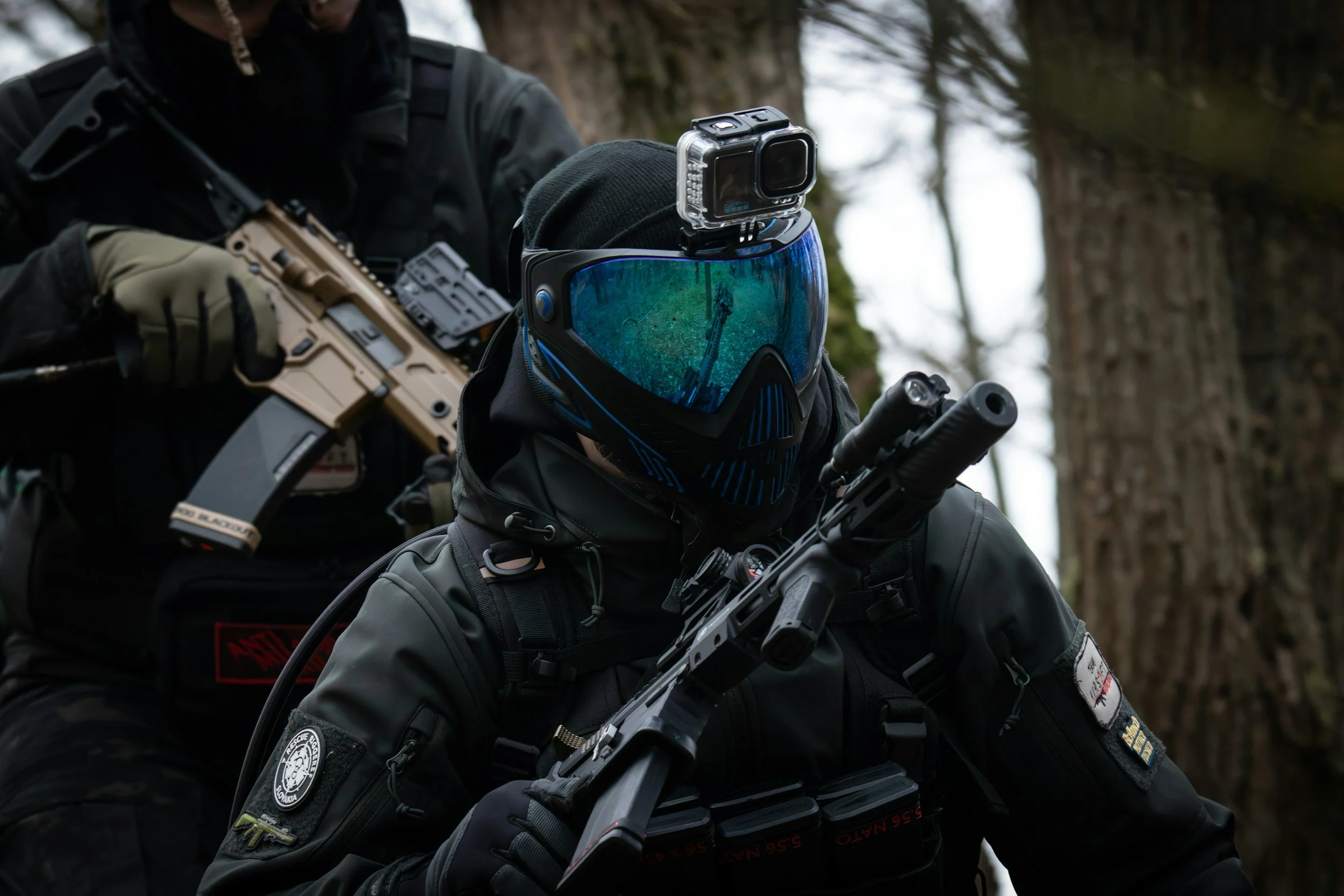 Two officers in tactical gear, one holding a firearm, stand outdoors near trees.