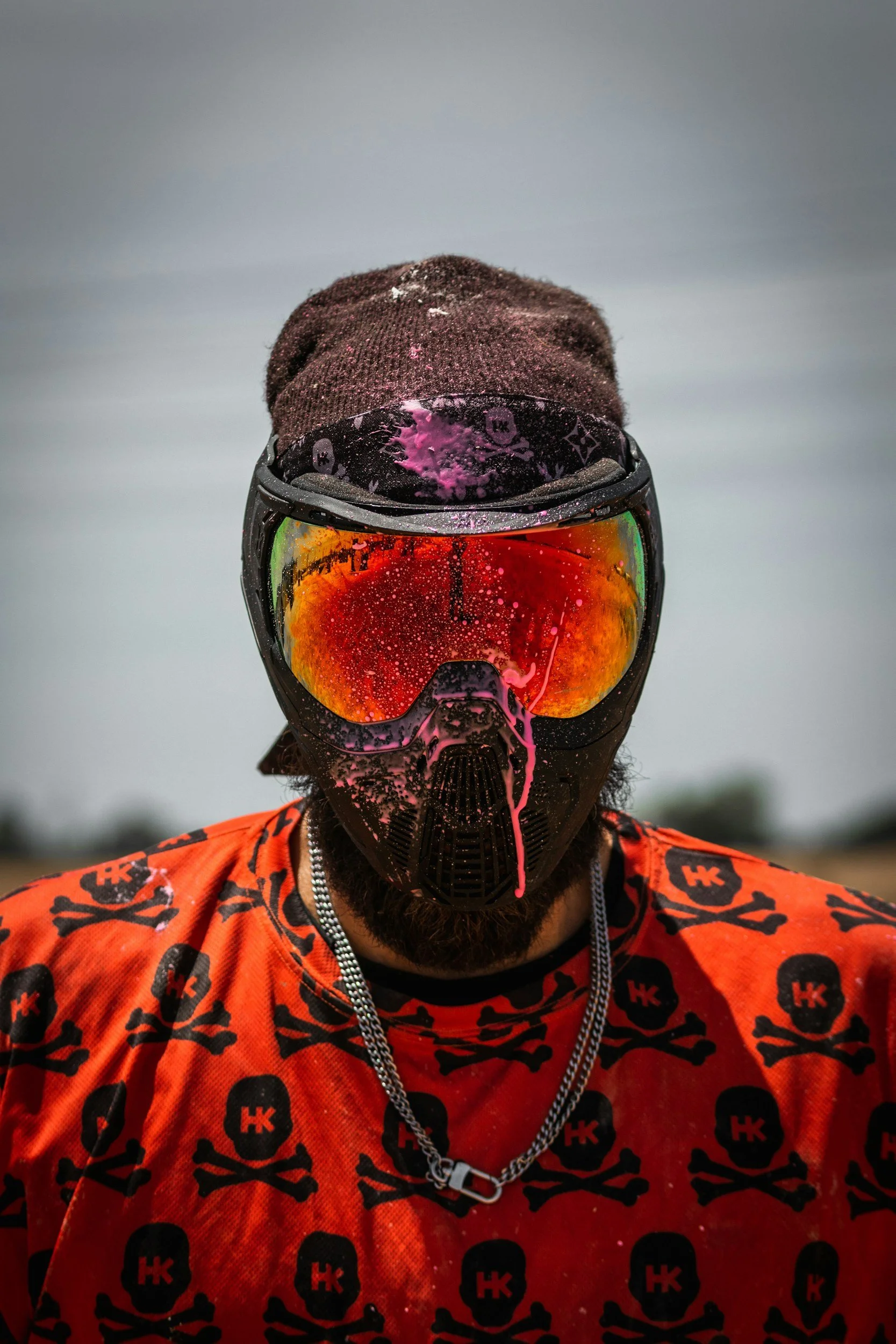 A person wearing a black helmet and goggles with orange reflective lenses, a brown knit cap, and an orange shirt with black skull and crossbones patterns. The helmet and goggles have pink paint splatters on them.