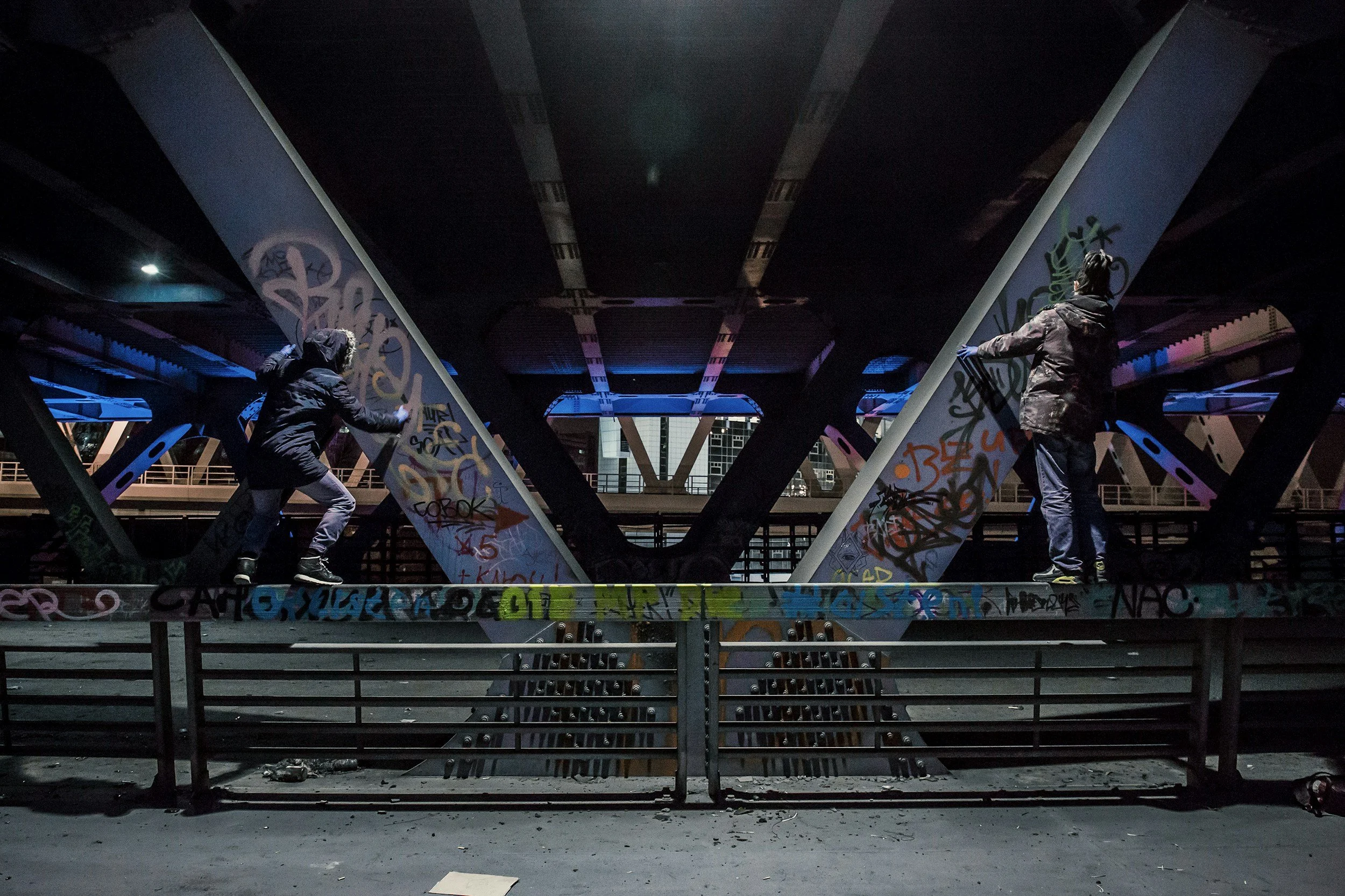 Two people spray-paint graffiti on urban structures beneath a bridge at night, with city buildings in the background.