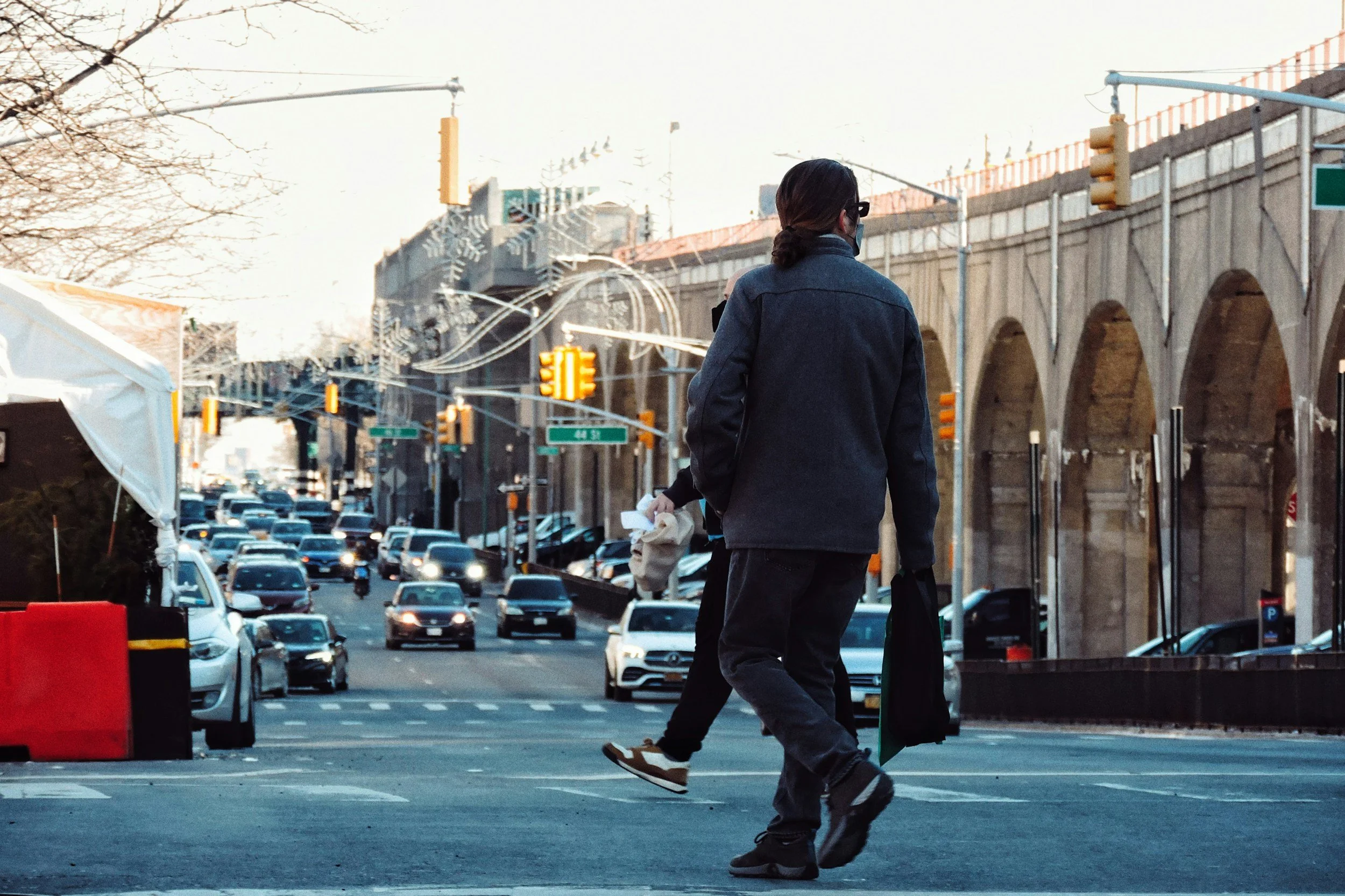 A man with long hair tied back, wearing a mask, sunglasses, a dark jacket, and jeans, is walking across a street in an urban area. There are traffic lights, cars, and a building with arches in the background, along with some trees and utility poles.