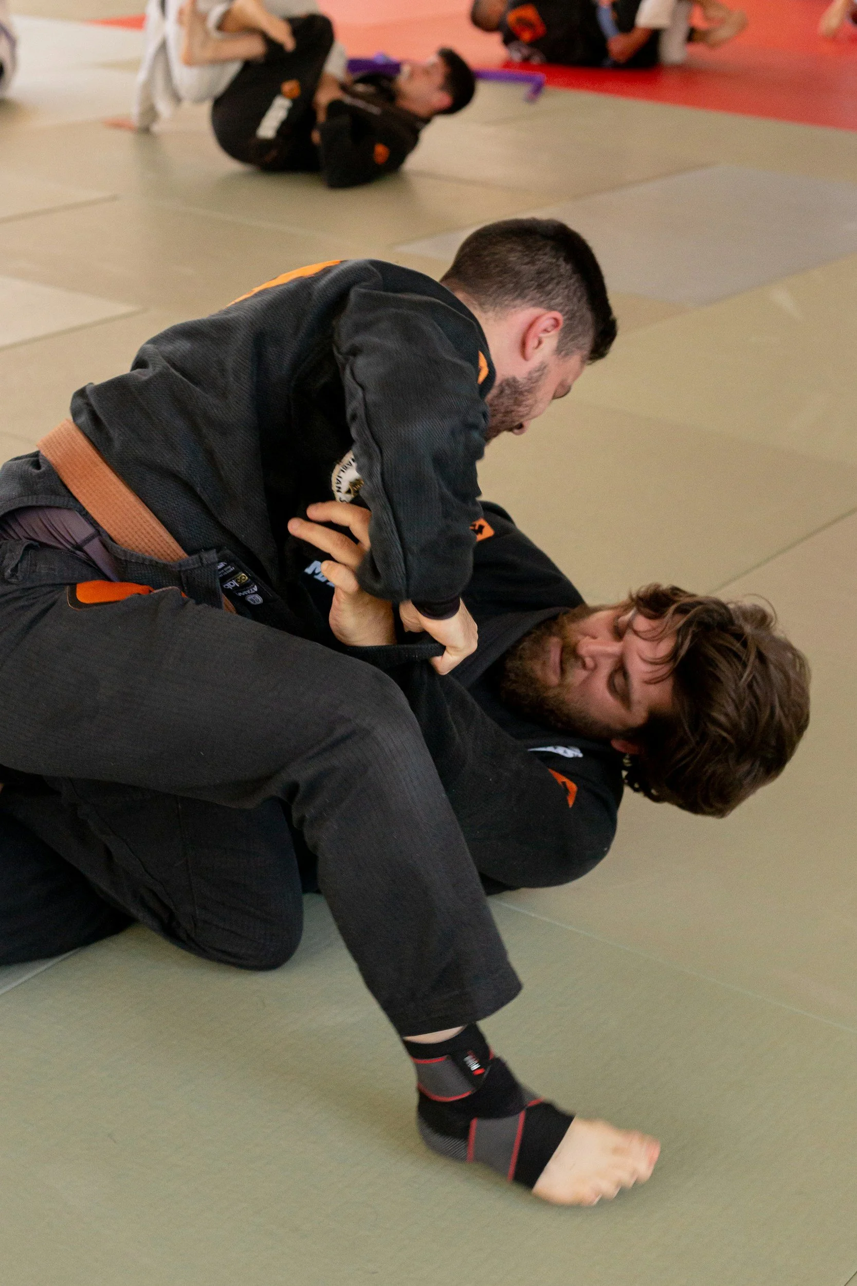Two men practicing Brazilian Jiu-Jitsu on a matted floor, one applying a submission hold while the other is on the ground in a defensive posture.
