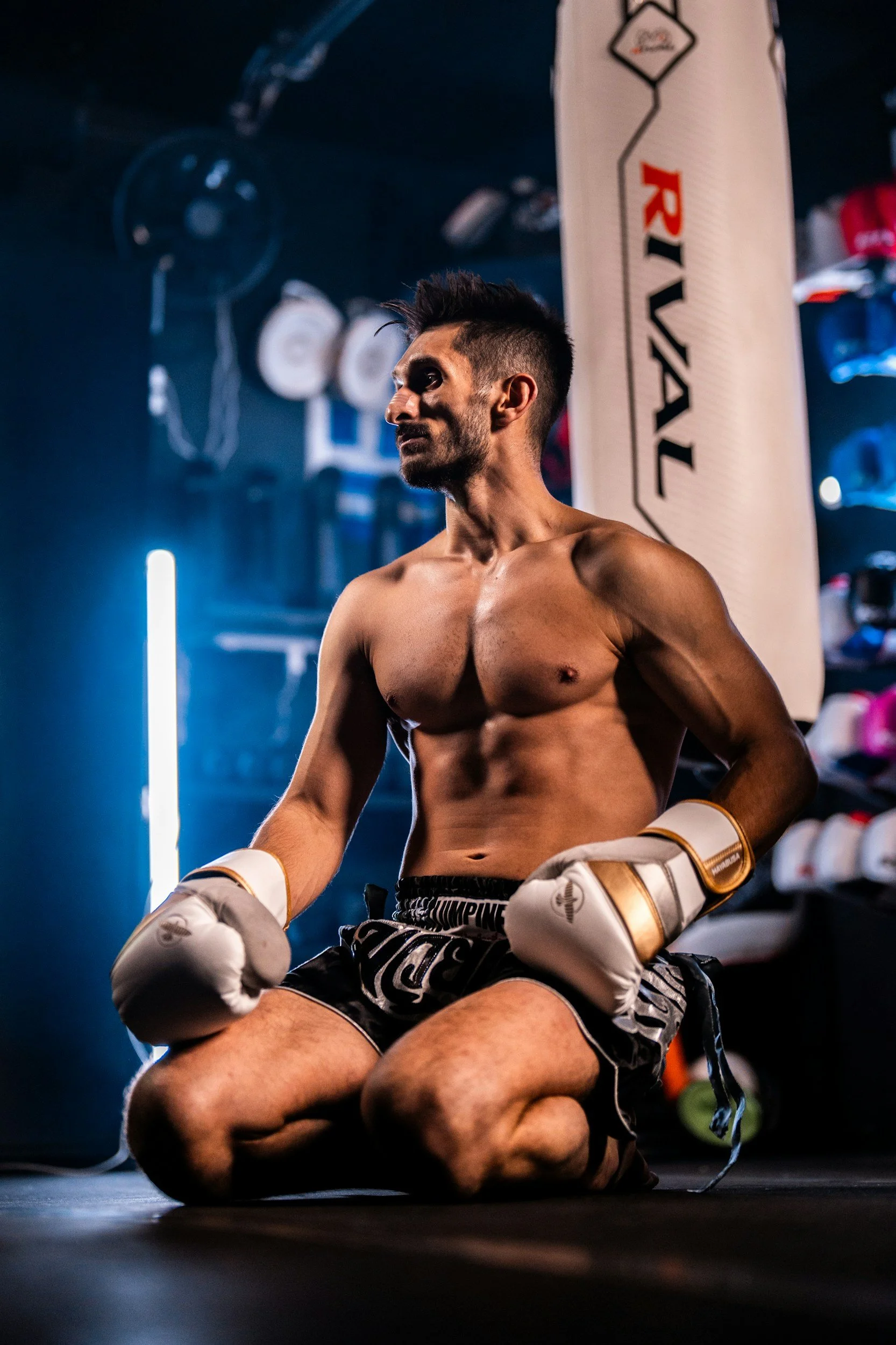 A shirtless male boxer wearing white gloves and black shorts, kneeling on the floor inside a boxing gym, with a large RIVAL punching bag in the background.
