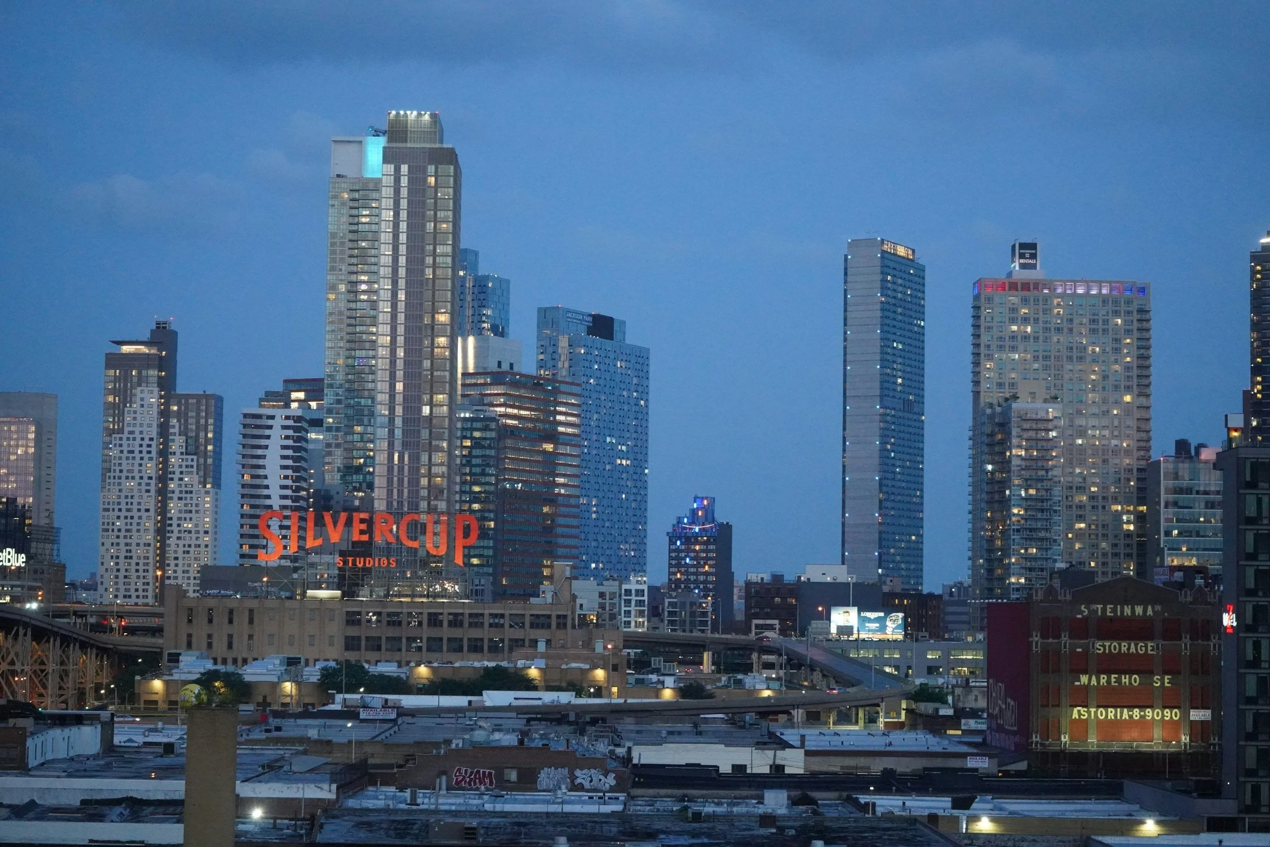 City skyline at dusk with tall modern buildings, including the Silvercup Studios sign, and a partly cloudy blue sky.