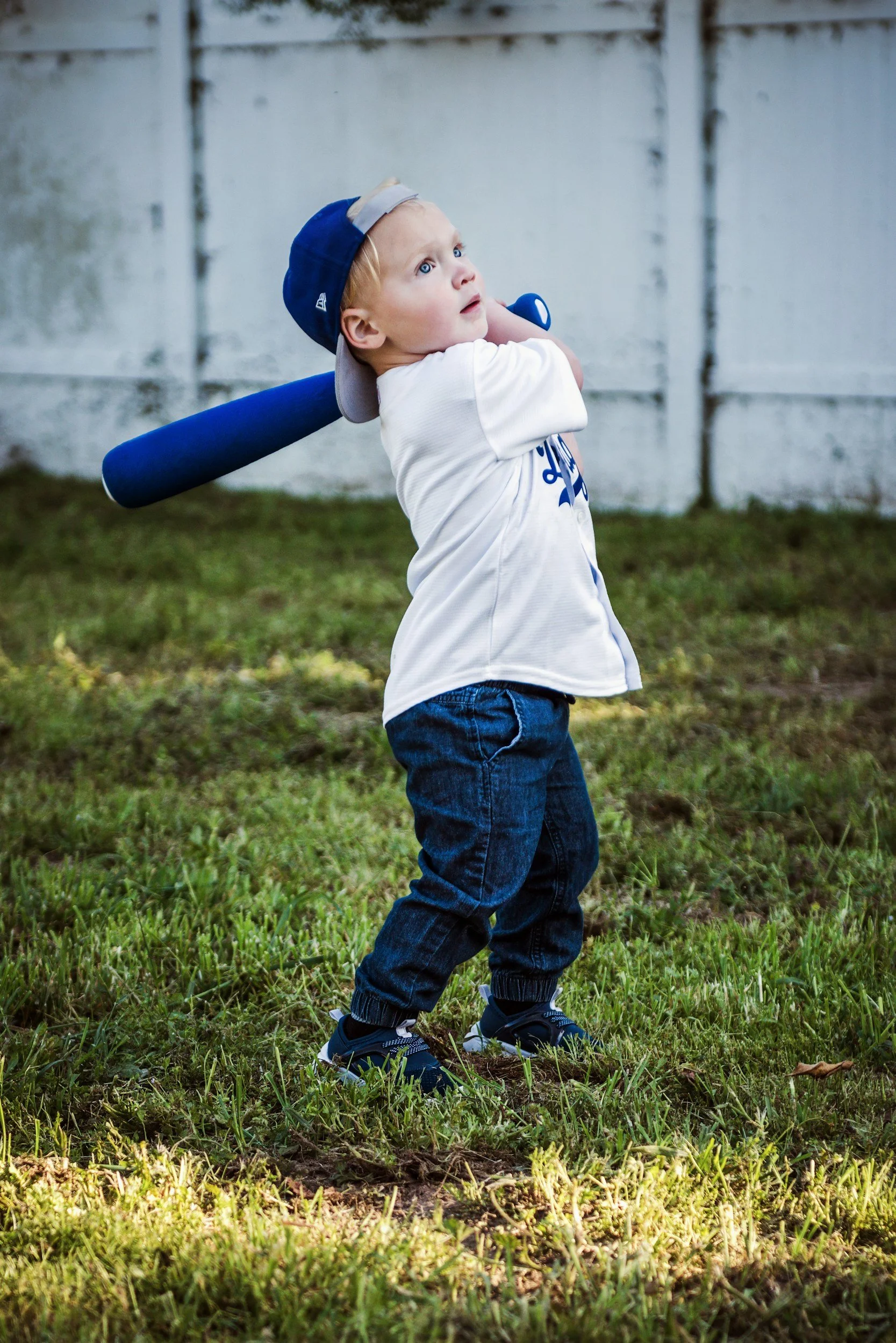 Young boy wearing a baseball cap backwards, holding a baseball bat, standing outdoors on grass, with a white fence in the background.