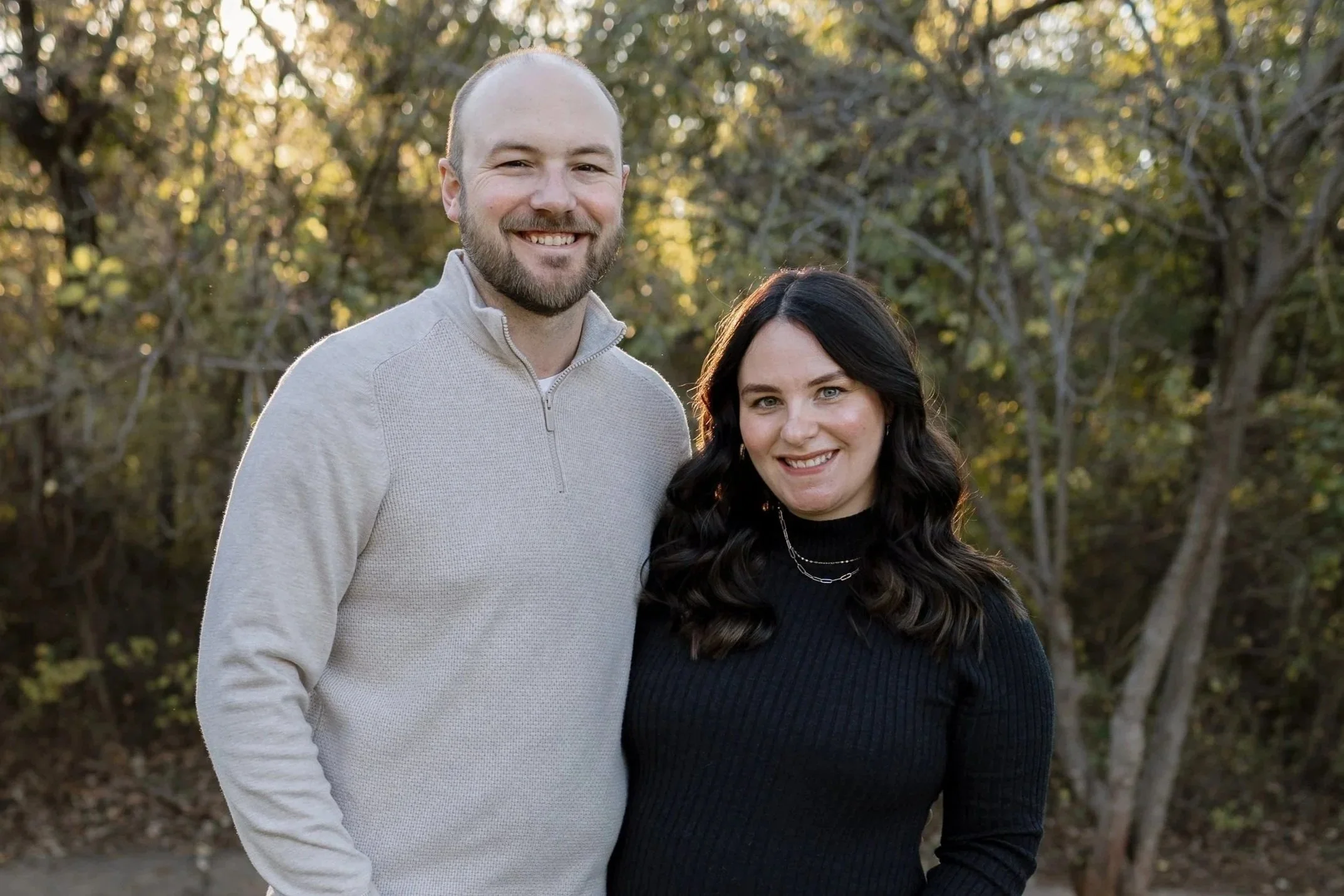 A smiling man with a beard and a woman with dark hair stand outdoors with trees in the background.