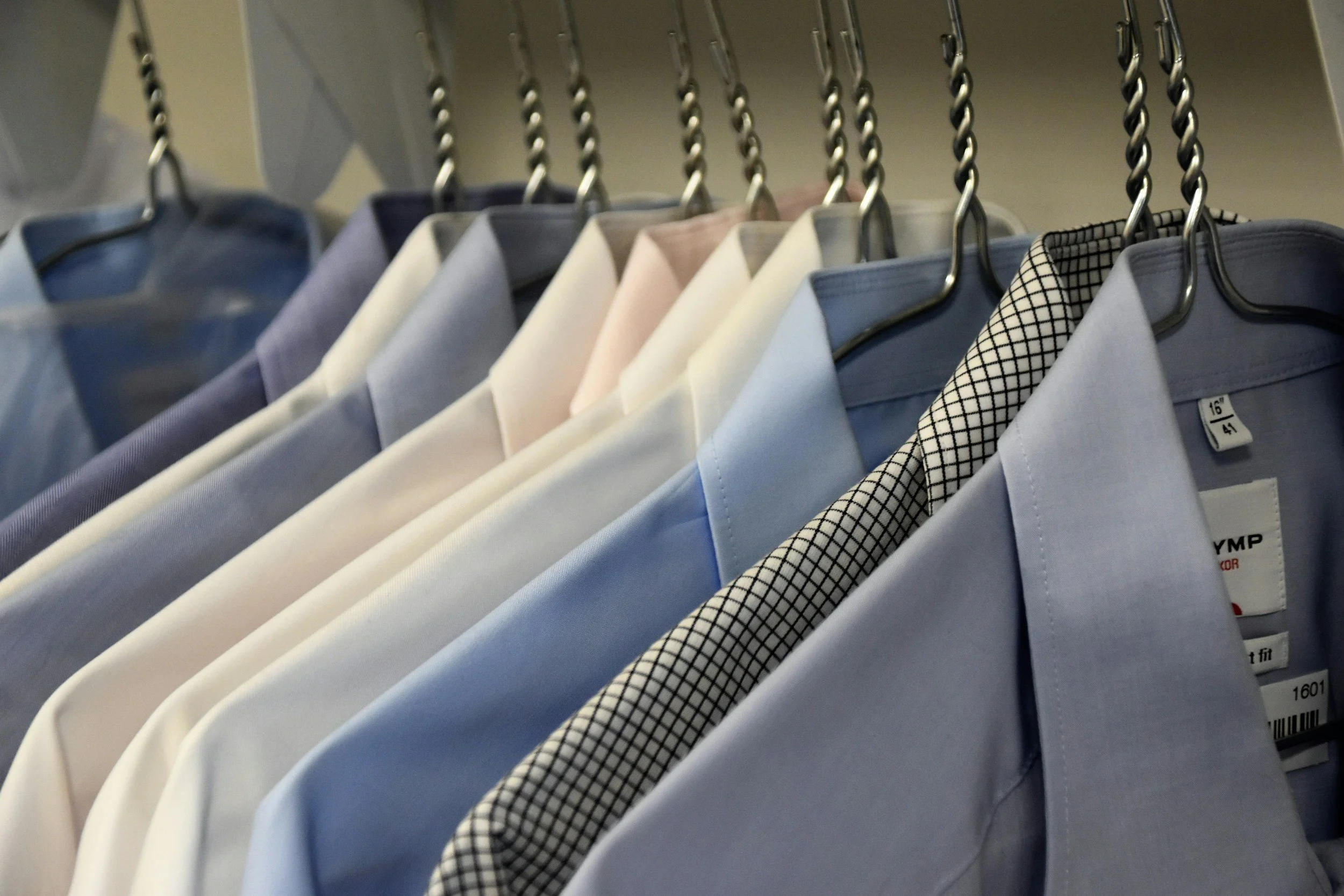 A row of men's dress shirts hanging on a clothing rack, with various colors and patterns, including solid light blue and pale pink, and a black and white checkered pattern.