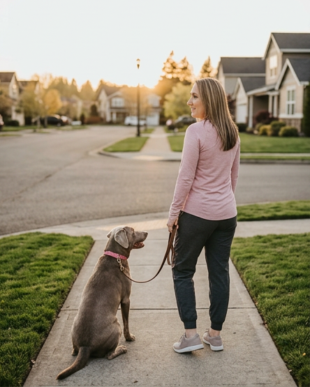 A woman walking her dog in a suburban neighborhood during sunset, with houses and a street in the background.