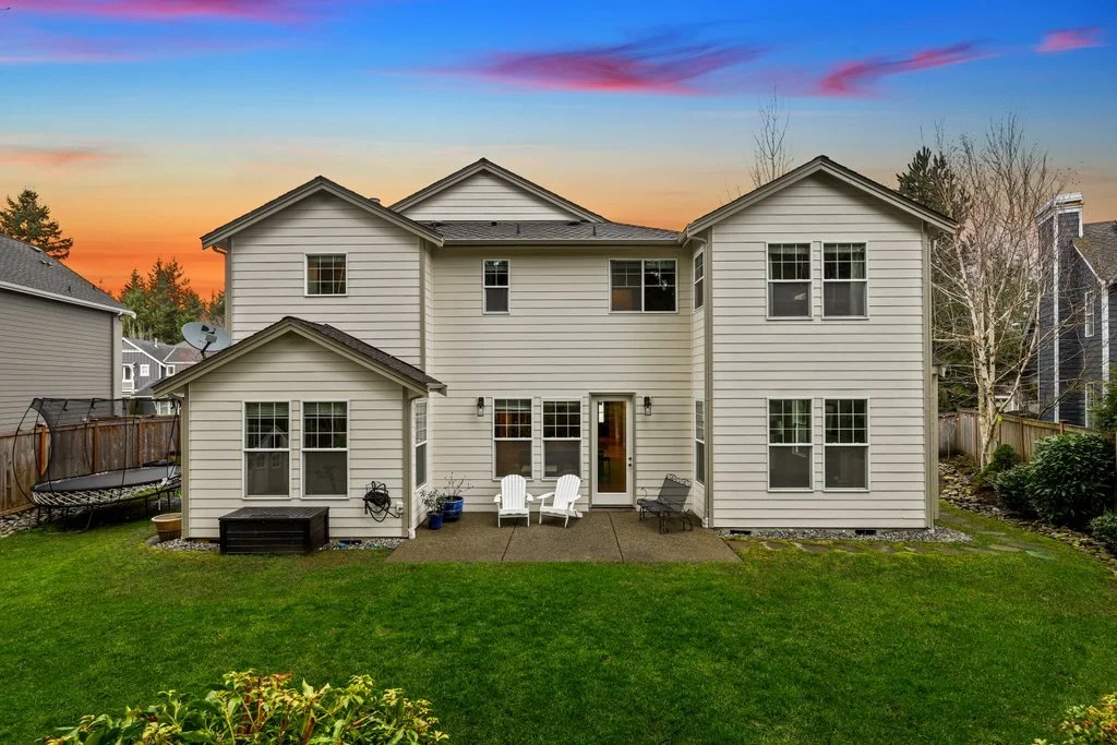 Backyard of a two-story house with beige siding during sunset, featuring a patio with white Adirondack chairs, a bench, and a black storage box, surrounded by a green lawn and neighboring houses.