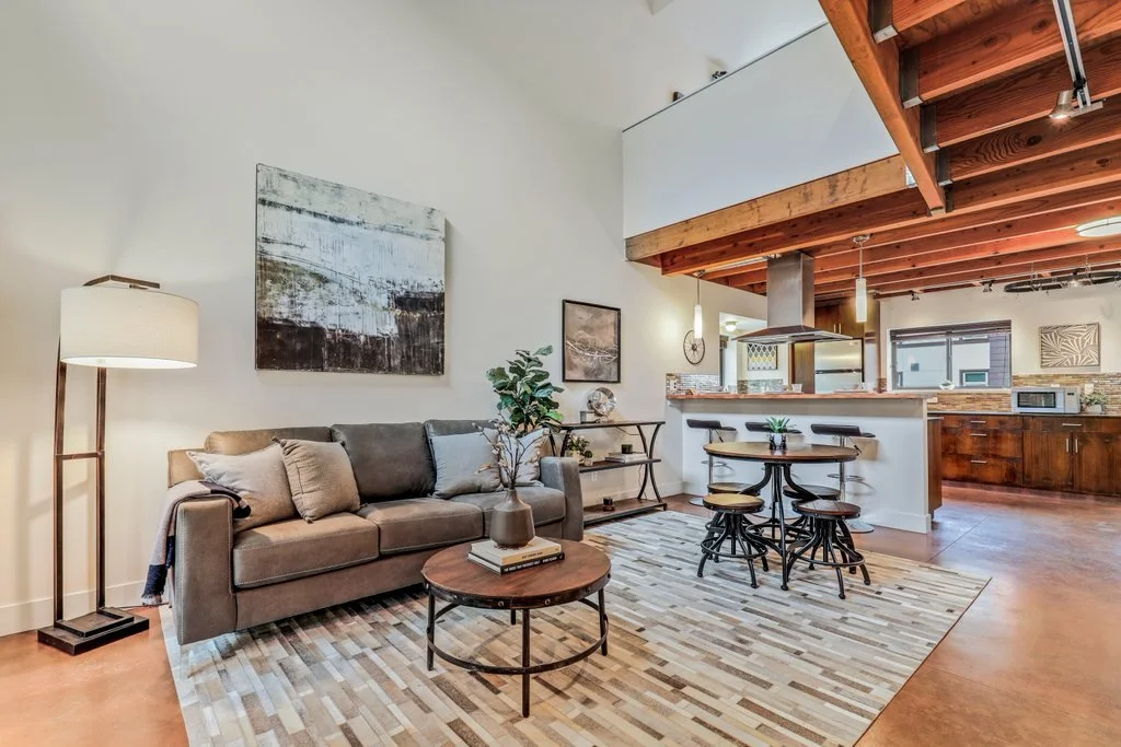 Modern living room with beige sofa, wooden coffee table, and abstract wall art, open kitchen with wood cabinets and stainless steel appliances, and a loft area above.