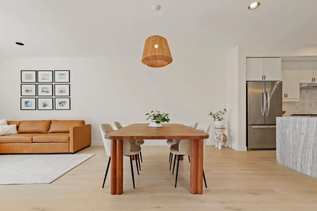 A modern open-plan living and dining area with a tan leather sofa, a light wood dining table with six white chairs, and a kitchen with a stainless steel refrigerator. The room has light-colored flooring, white walls, and minimalistic decor, including