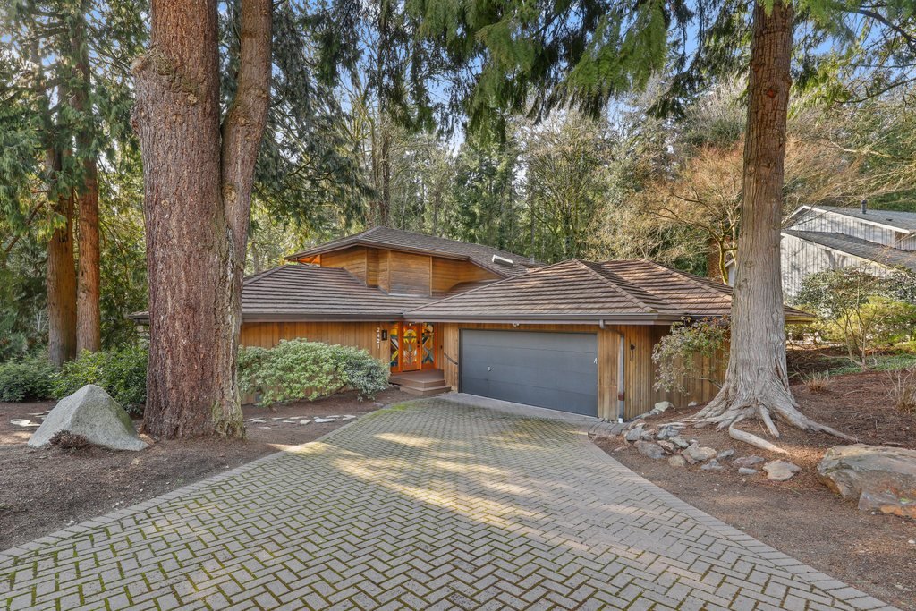 A wooden house with a garage, surrounded by trees and greenery, with a paved driveway leading up to it.