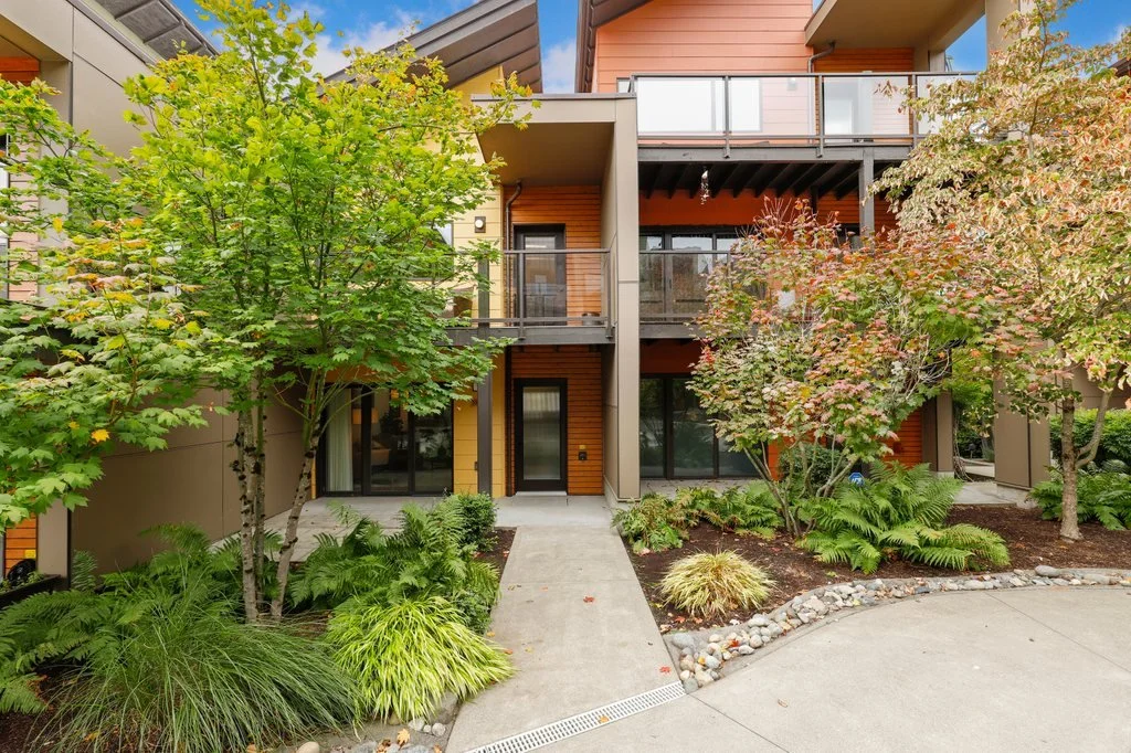 Front view of a modern apartment building with balconies, surrounded by lush green trees and landscaped garden.