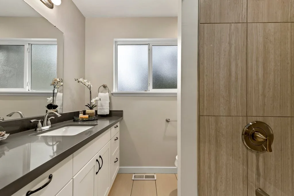A modern bathroom with a large mirror above a white vanity with black handles, a gray countertop, a built-in sink, and decorative plants, towels, and toiletries. There are two frosted windows and a wooden-paneled wall with a bronze towel hook.