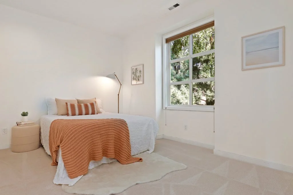 A minimalistic bedroom with a white bed, beige pillows, a rust-colored throw blanket, a small nightstand with a plant and books, a wall-mounted reading lamp, a large window with greenery outside, and two framed pictures on the wall.