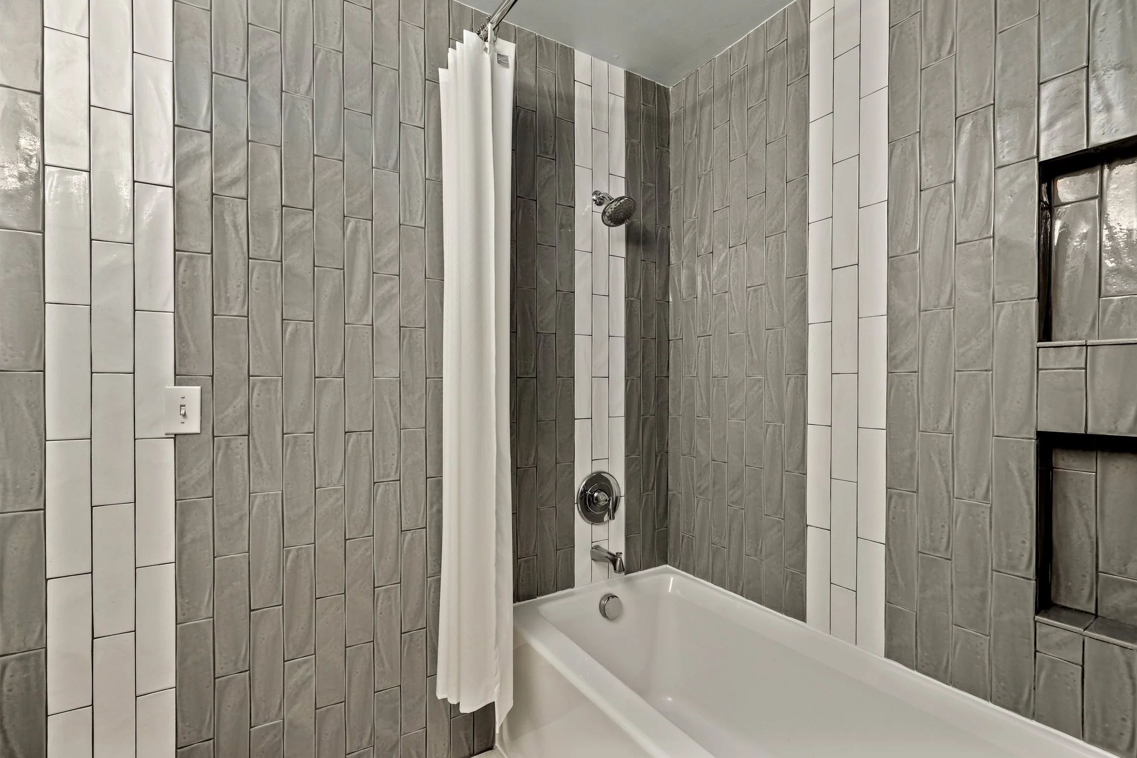Bathroom shower with gray and white tiles, a white bathtub, a showerhead, a shower curtain, and an opening in the wall.