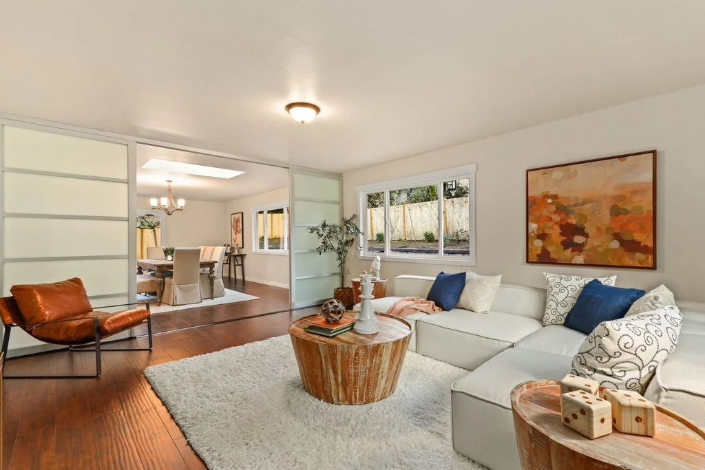 Living room with cream sectional couch, decorative pillows, wooden coffee table, area rug, and wall art, with an adjacent dining area visible through a sliding door.