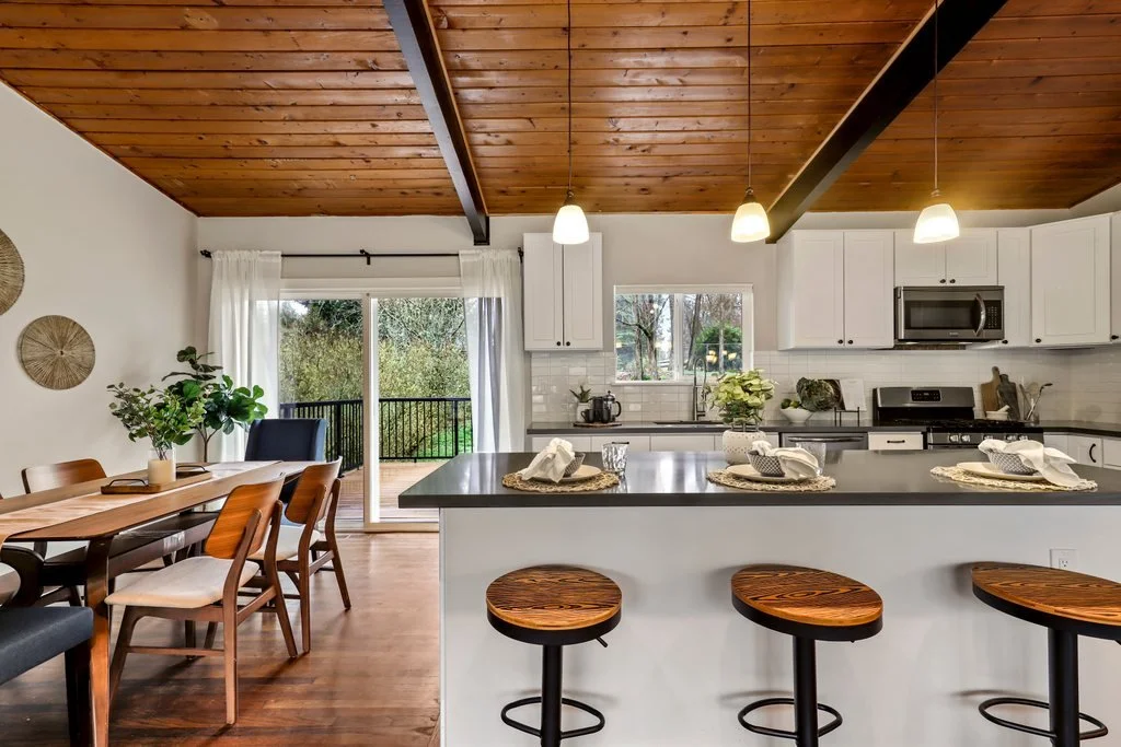 Open-concept kitchen and dining area with wooden ceiling, white cabinetry, and a sliding glass door leading to an outdoor deck with trees.