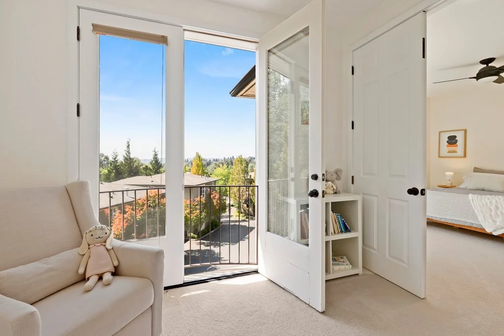 A bright bedroom with an open door leading to a balcony with a view of trees, rooftops, and a blue sky, and a plush doll on a cream-colored armchair.