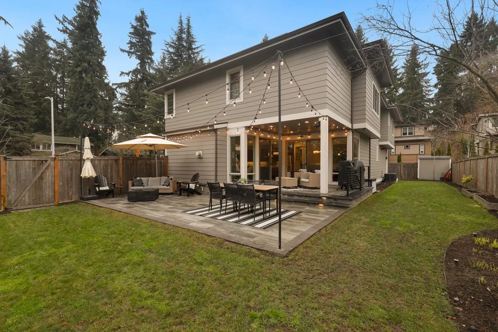 Backyard patio of a house with outdoor furniture, string lights, a large umbrella, and a grassy yard surrounded by a wooden fence, with trees in the background.