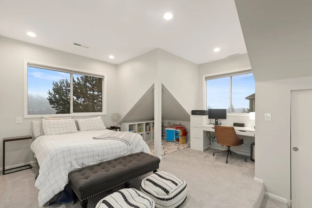 A bedroom with a bed, two windows, a small bookshelf with toys, a desk with a computer, and two ottomans, in a bright space with white walls and ceiling lights.
