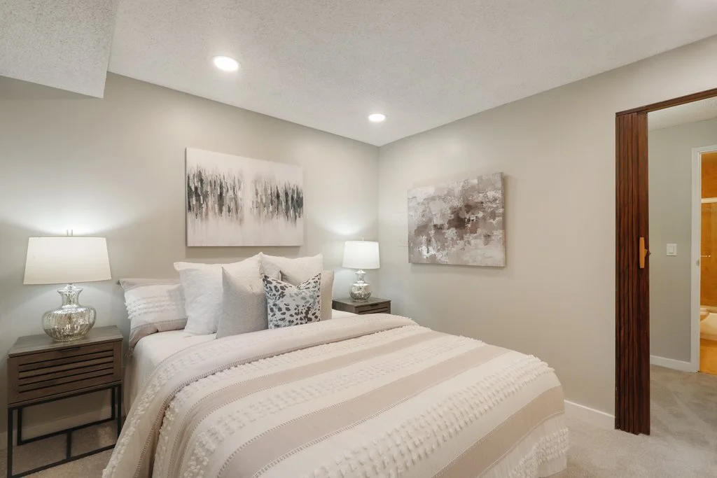 A bedroom with a bed, pillows, and artwork on the walls, illuminated by two table lamps and ceiling lights.