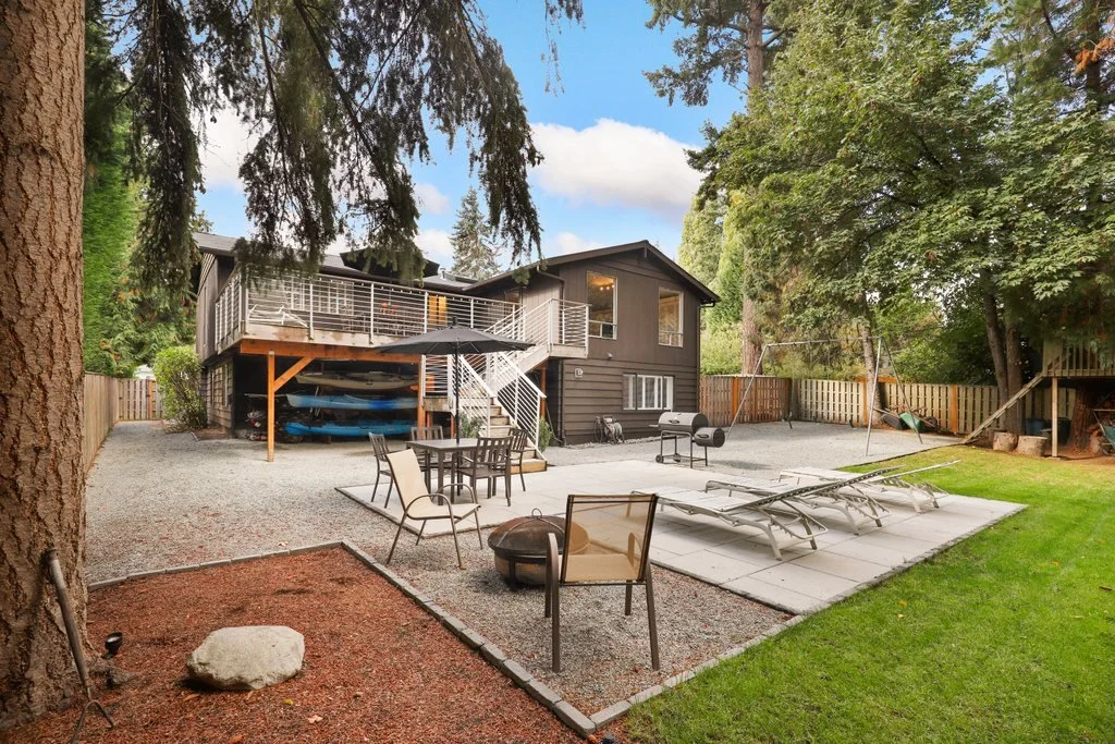 Backyard with a raised deck, outdoor furniture, lounge chairs, a grill, a swing set, and a grassy area, surrounded by a wooden fence and tall trees.