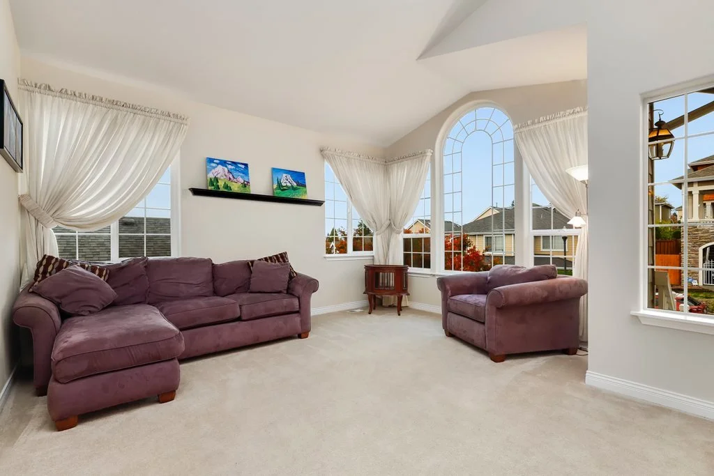 Living room with purple sofas, large windows with white curtains, and a small wooden table.