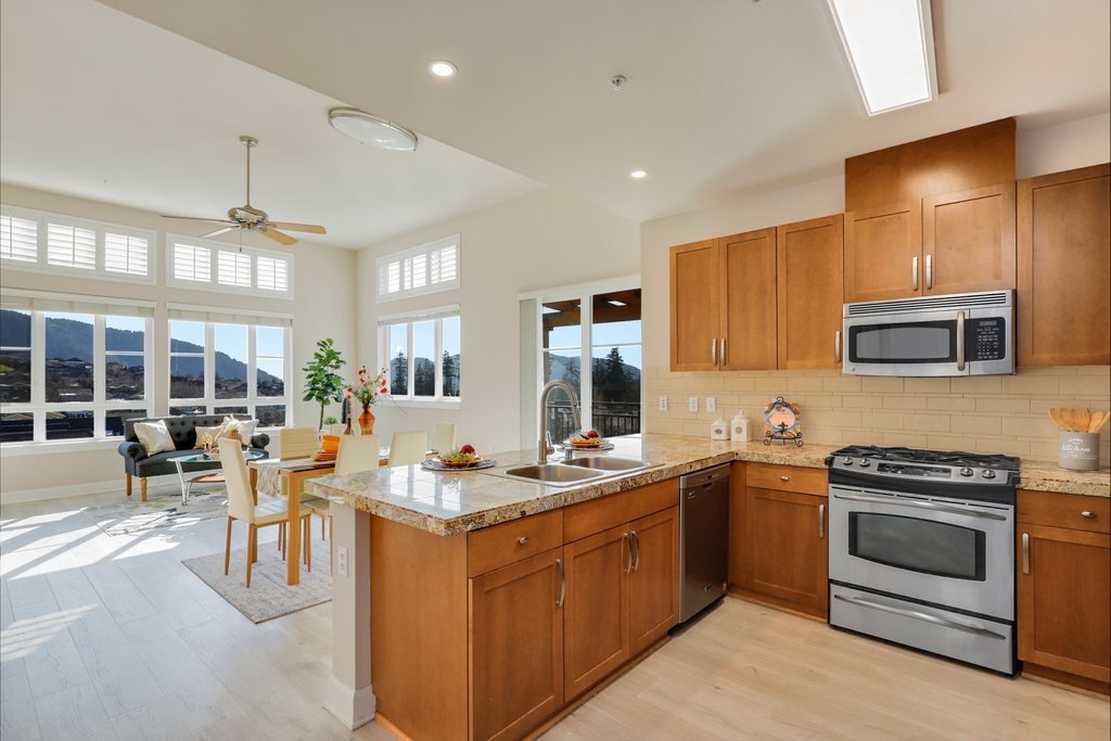 Open-concept living room and kitchen with large windows, wooden cabinets, granite countertops, stainless steel appliances, dining table, and a sitting area with a black couch and chairs.