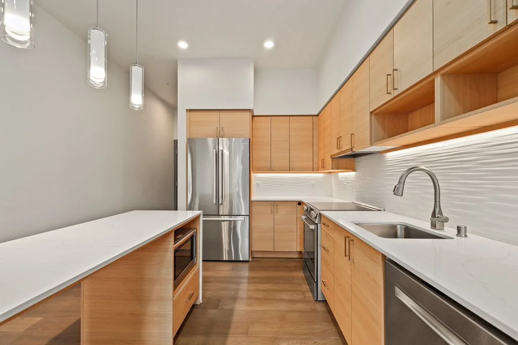 Modern kitchen with light wood cabinets, stainless steel refrigerator, stove, dishwasher, and white countertops with sleek gray backsplash.