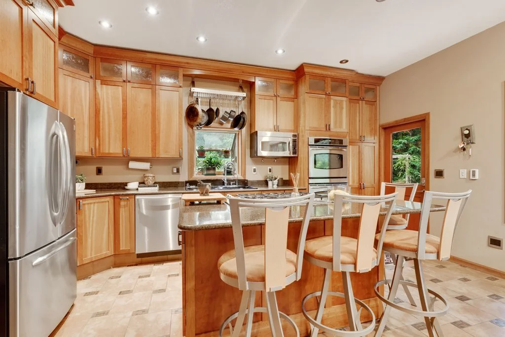 Bright kitchen with wooden cabinets, granite countertops, stainless steel appliances, and a breakfast bar with four white chairs.