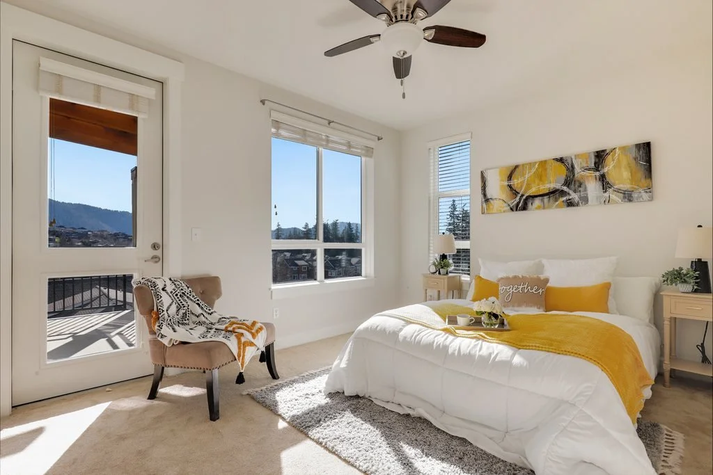 Bright bedroom with a large bed featuring white bedding, yellow accents, and a decorative pillow saying 'together'. Two bedside tables with white lamps, a beige chair with a black and white throw blanket, a large window with a view of mountains and t