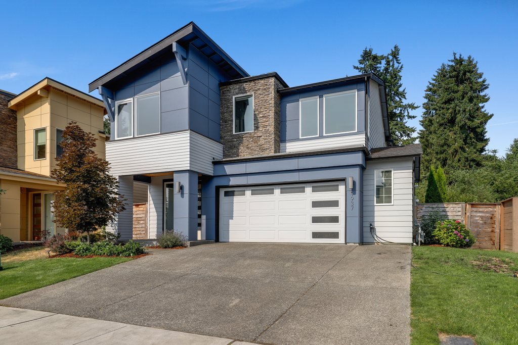 Modern two-story house with a garage, flat roof, large windows, and a landscaped front yard.