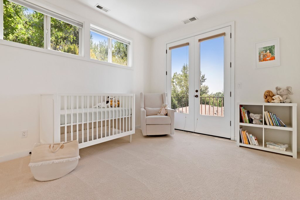 A nursery with white walls and furniture, including a crib with stuffed toys, a beige armchair with a stuffed animal, a small bookshelf with books and plush toys, and a glass door leading outside, with a view of trees and rooftops.
