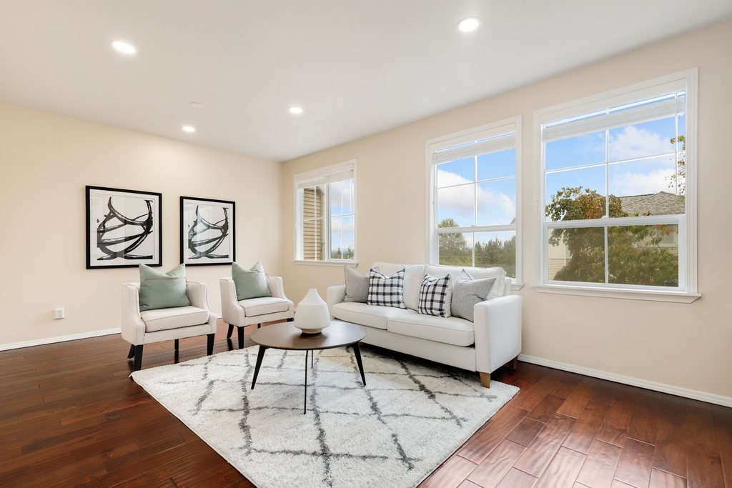 Bright living room with white sofa, two white armchairs with light green pillows, and a round coffee table on a white and gray patterned rug. There are three black-and-white framed pictures on the wall and three windows letting in natural light.