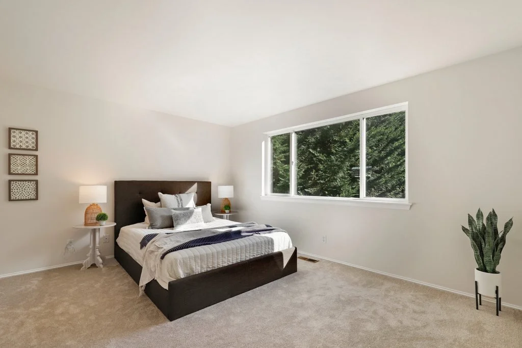 A minimalistic bedroom with a bed, two bedside tables with lamps, a window with trees outside, and a potted plant on the floor.