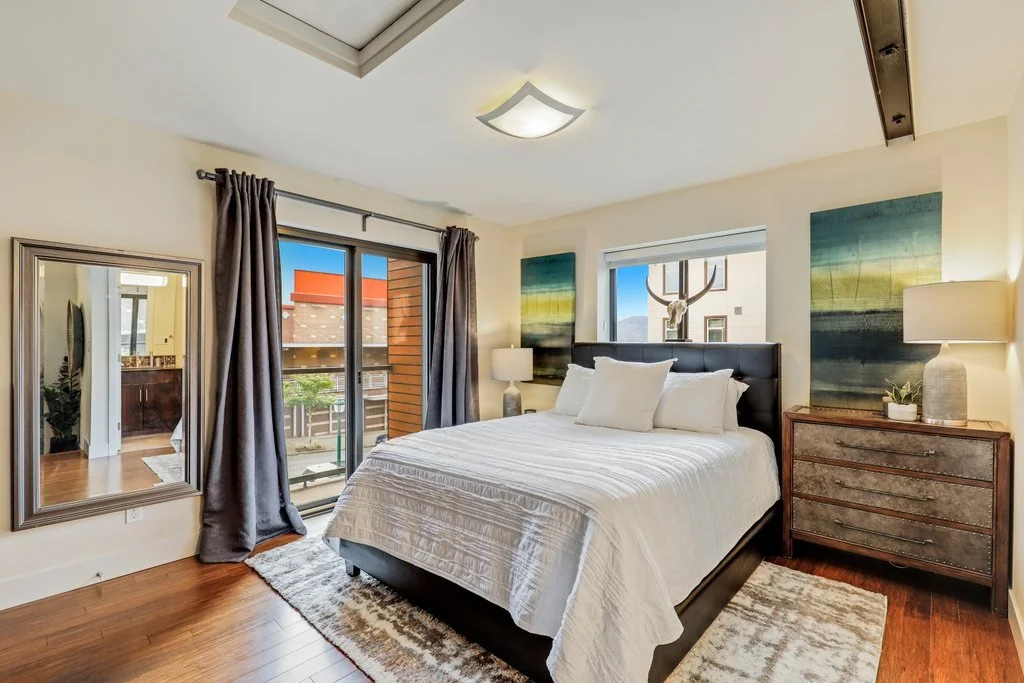 A bedroom with a large bed, dark headboard, white bedding and pillows, wooden nightstand with lamp, artwork on walls, sliding glass door with curtains leading to a small balcony, and another window behind the bed with a decorative deer head mounted o