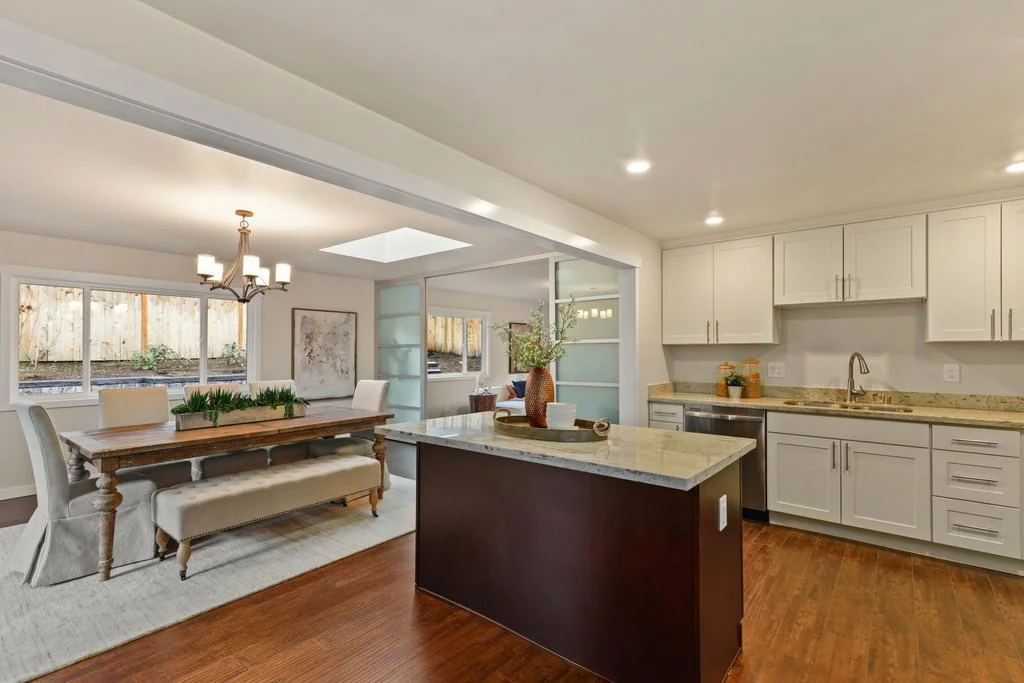 Open-concept kitchen and dining area with white cabinets, a kitchen island, a dining table with chairs, and large windows providing natural light.