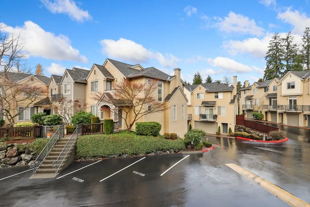 Residential apartment complex with multiple beige buildings, landscaped greenery, wet parking lot, and clear sky with clouds.