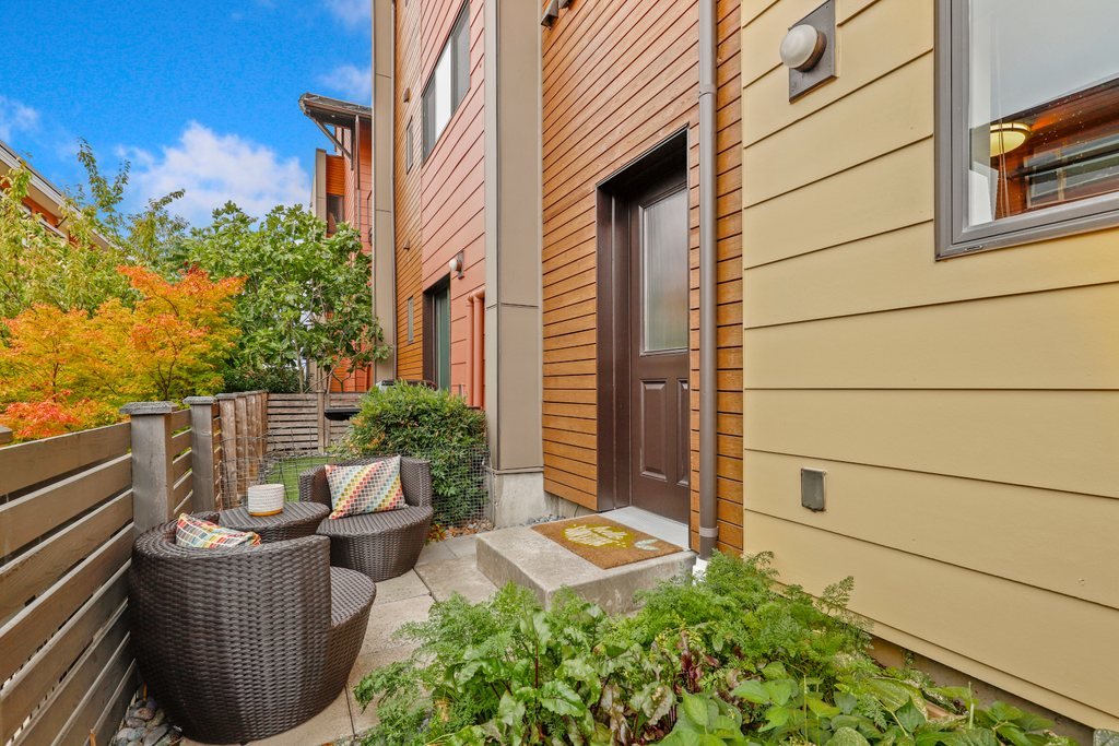 Small outdoor patio area with two wicker chairs, colorful cushions, a small table, and plants with colorful trees in the background, next to a building with tan and wooden exterior walls.