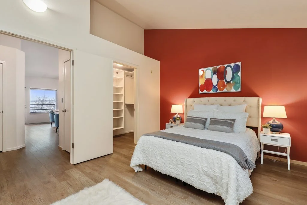 A bedroom with a red accent wall, a cream tufted headboard, white bedding, two lamps on white nightstands, and a colorful abstract painting above the bed.
