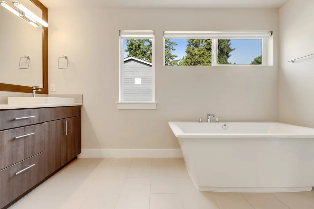 A modern bathroom with a wooden vanity, a white countertop, a sink, a mirror, a large bathtub, and windows with a view of trees outside.